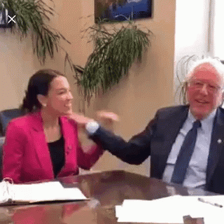 A Man And A Woman Are Sitting At A Table Shaking Hands . The Woman Is Wearing A Pink Jacket . GIF
