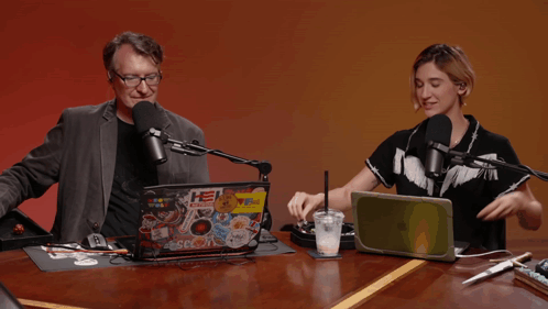 A Man And A Woman Are Sitting At A Table With A Laptop With Stickers On It That Says He Radio GIF
