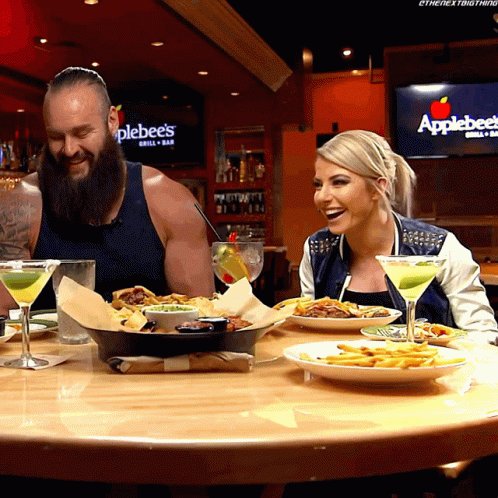 A Man And A Woman Are Sitting At A Table With Plates Of Food In Front Of An Applebee 'S Sign GIF