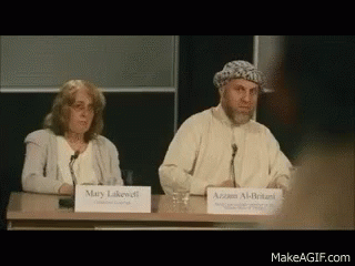 A Man And A Woman Are Sitting At A Table With Signs That Say Mary Lakewell And Azzam Al-Britanni GIF