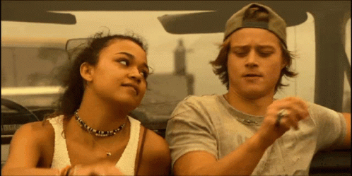 A Man And A Woman Are Sitting In A Car And The Woman Is Wearing A Necklace GIF