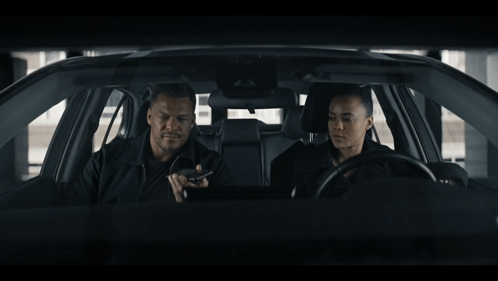 A Man And A Woman Are Sitting In A Car Looking At Something GIF