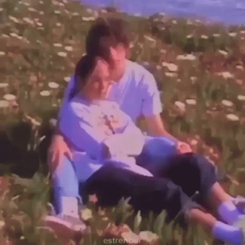 A Man And A Woman Are Sitting In A Field Of Flowers And Hugging Each Other . GIF