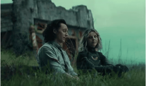 A Man And A Woman Are Sitting In The Grass Next To A Building . GIF