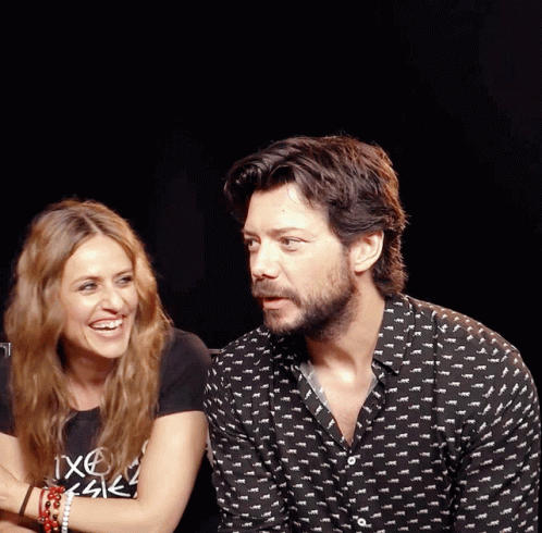 A Man And A Woman Are Sitting Next To Each Other And The Man Is Wearing A Shirt That Says " Ixcel " On It GIF