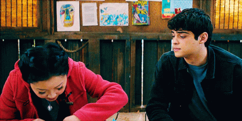 A Man And A Woman Are Sitting Next To Each Other In Front Of A Bulletin Board GIF