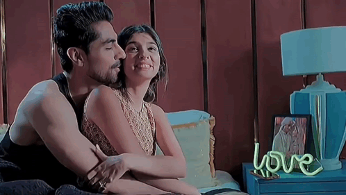 A Man And A Woman Are Sitting On A Bed With A Neon Sign That Says Love On The Nightstand . GIF