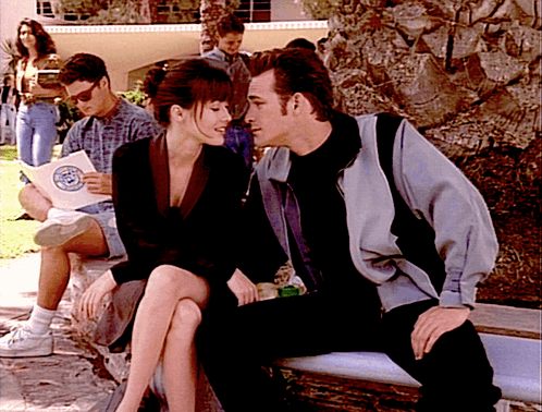A Man And A Woman Are Sitting On A Bench And The Man Is Reading A Book With A Circle On It GIF