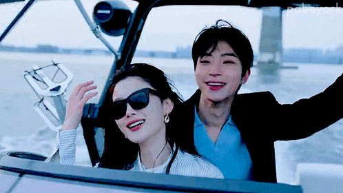 A Man And A Woman Are Sitting On A Boat And The Man Is Wearing Sunglasses And The Woman Is Wearing Earrings GIF