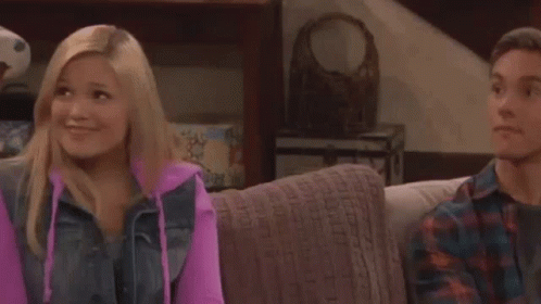 A Man And A Woman Are Sitting On A Couch And Smiling . The Woman Is Wearing A Pink Hoodie . GIF