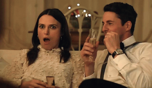 A Man And A Woman Are Sitting On A Couch Holding Glasses Of Champagne GIF