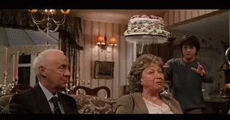 A Man And A Woman Are Sitting On A Couch In A Living Room With A Cake On Their Head . GIF