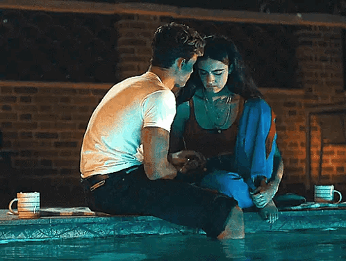 A Man And A Woman Are Sitting On The Edge Of A Swimming Pool Looking At Each Other . GIF