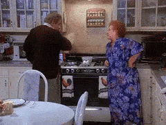 A Man And A Woman Are Standing In A Kitchen Looking At A Stove Top Oven GIF