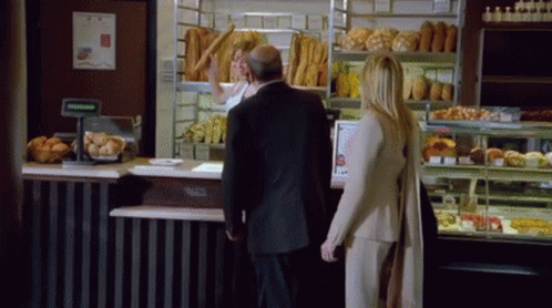 A Man And A Woman Are Standing In Front Of A Bakery GIF