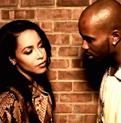 A Man And A Woman Are Standing In Front Of A Brick Wall And Looking At Each Other GIF