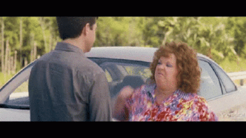 A Man And A Woman Are Standing In Front Of A Car And Talking To Each Other . GIF