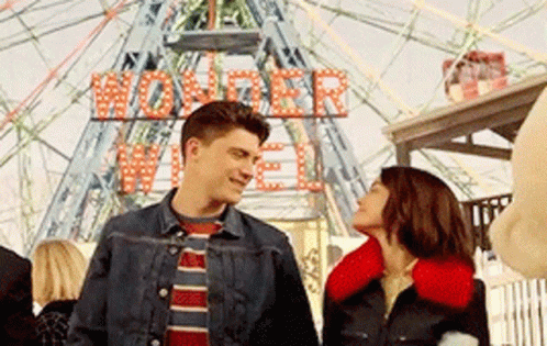 A Man And A Woman Are Standing In Front Of A Ferris Wheel That Says Wonder Wheel On It GIF