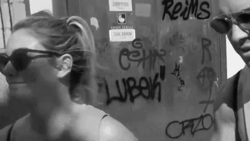A Man And A Woman Are Standing In Front Of A Wall With Graffiti On It Including Reims GIF