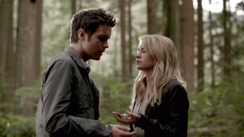 A Man And A Woman Are Standing In The Woods And The Man Is Holding Something In His Hand GIF