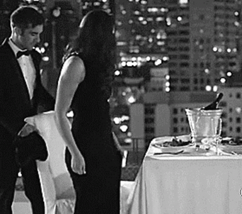 A Man And A Woman Are Standing Next To A Table With A Bottle Of Champagne . GIF
