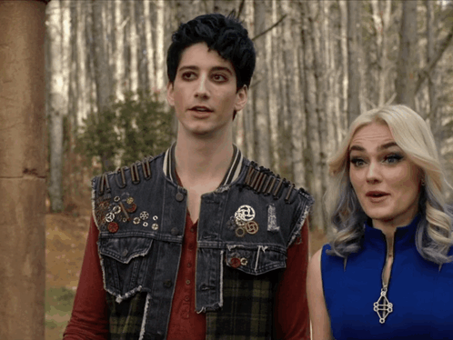 A Man And A Woman Are Standing Next To Each Other And The Man Is Wearing A Vest With Pins On It GIF