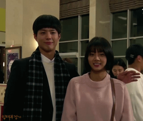 A Man And A Woman Are Standing Next To Each Other And The Woman Is Wearing A Pink Sweater And Scarf GIF