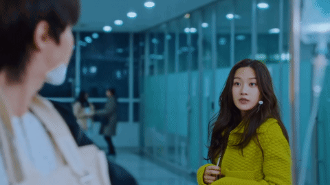 A Man And A Woman Are Standing Next To Each Other In A Hallway . The Woman Is Wearing A Green Sweater . GIF