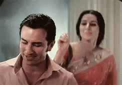 A Man And A Woman Are Standing Next To Each Other In A Room . The Woman Is Holding The Man 'S Hair . GIF