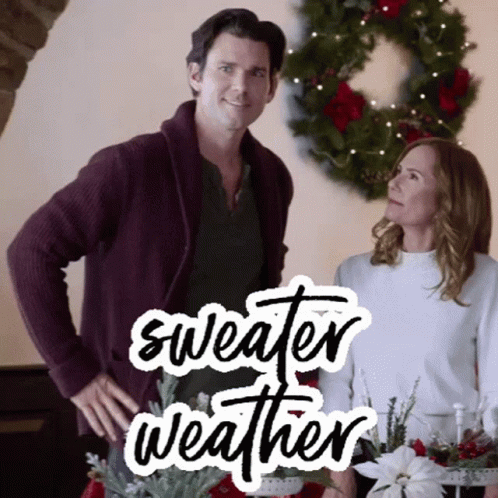 A Man And A Woman Are Standing Next To Each Other In Front Of A Christmas Wreath With The Words Sweater Weather Written GIF
