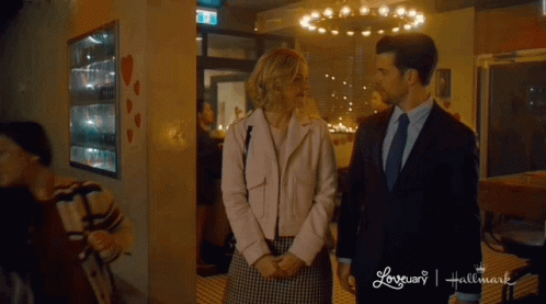 A Man And A Woman Are Standing Next To Each Other In Front Of A Hallmark Sign GIF