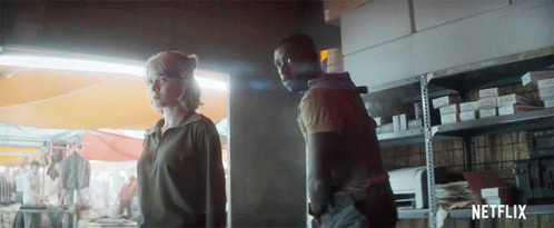 A Man And A Woman Are Standing Next To Each Other In Front Of A Shelf With Netflix Written On It GIF