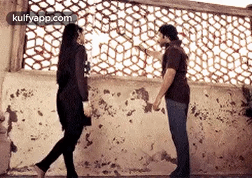 A Man And A Woman Are Standing Next To Each Other In Front Of A Wall Holding Hands . GIF