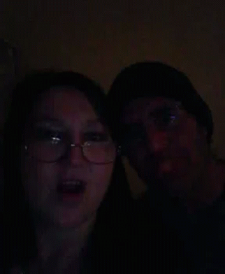A Man And A Woman Are Standing Next To Each Other In The Dark . The Woman Is Wearing Glasses And A Hat . GIF