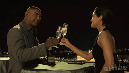 A Man And A Woman Are Toasting With Champagne And The Killer 'S Game Is Visible In The Background GIF