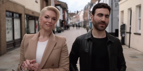 A Man And A Woman Are Walking Down A Street And The Woman Is Wearing Earrings GIF