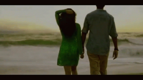 A Man And A Woman Are Walking On The Beach Holding Hands . The Woman Is Wearing A Green Dress . GIF