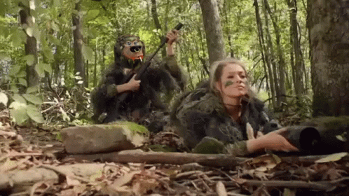 A Man And A Woman In Camouflage Are Laying In The Woods . GIF