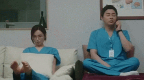 A Man And A Woman In Scrubs Are Sitting On A Couch With Their Feet Crossed . GIF