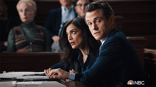 A Man And A Woman Sit At A Table In A Courtroom With #Lawandorder On The Bottom Right GIF