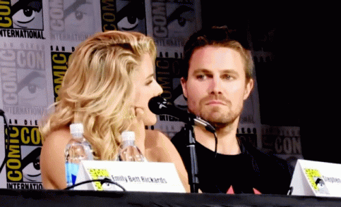 A Man And A Woman Sit At A Table With A Comic Con Sign Behind Them GIF