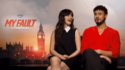 A Man And A Woman Sit Next To Each Other In Front Of A Poster For My Fault London GIF