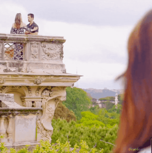 A Man And A Woman Standing On A Balcony Overlooking A Lush Green Forest With A Watermark That Says Corteco GIF