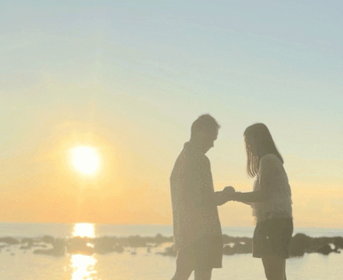 A Man And A Woman Standing On A Beach Looking At The Sunset GIF