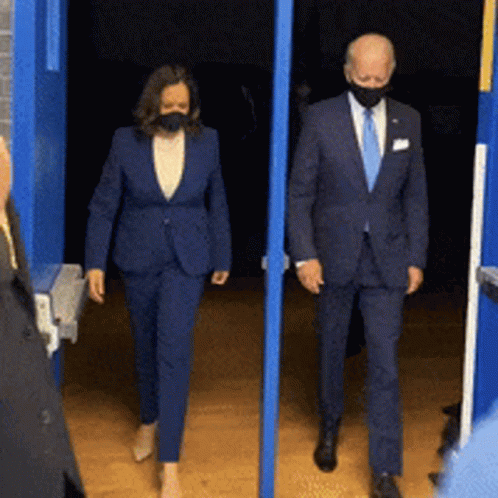 A Man And A Woman Wearing Masks Are Walking Out Of A Doorway GIF