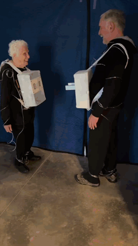 A Man And An Older Woman Are Standing Next To Each Other Wearing Cardboard Boxes On Their Arms GIF