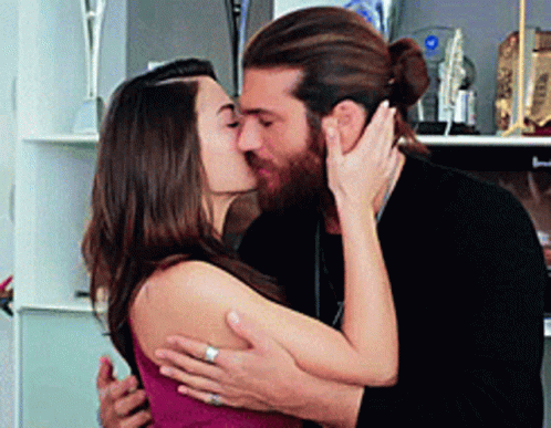 A Man And Woman Are Kissing In Front Of A Shelf With Trophies On It . GIF