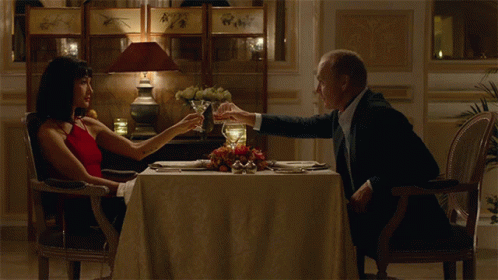 A Man And Woman Are Sitting At A Table With Glasses Of Wine GIF