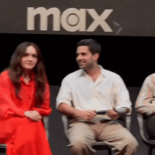 A Man And Woman Are Sitting Next To Each Other In Front Of A Max Sign . GIF