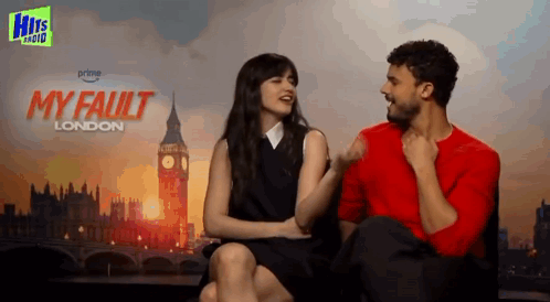 A Man And Woman Are Sitting Next To Each Other In Front Of A Poster For My Fault London GIF